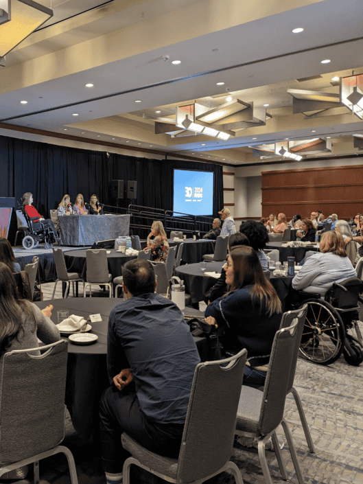 Photo of a conference room full of people. To the left is a stage showing Leah moderating, along with three panelists, seated.